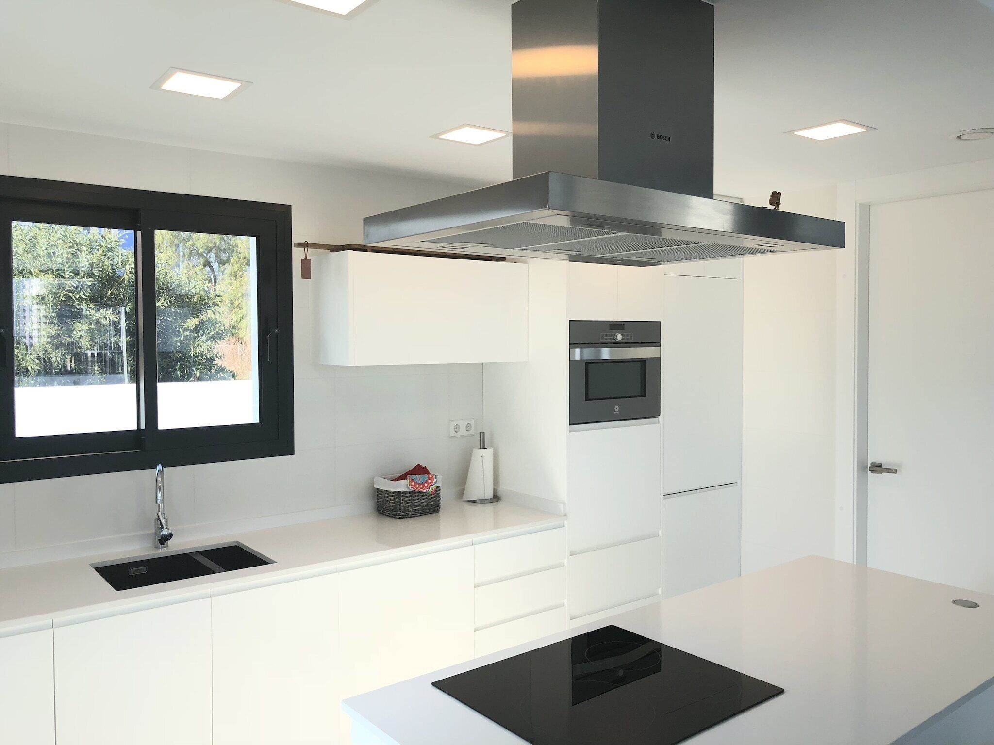 Une cuisine moderne avec des meubles blancs, un four encastré et une fenêtre donnant sur un paysage verdoyant.