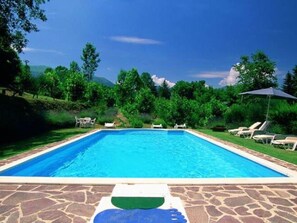 Piscina rectangular con borde de piedra, rodeada de césped, árboles y cielo azul despejado.