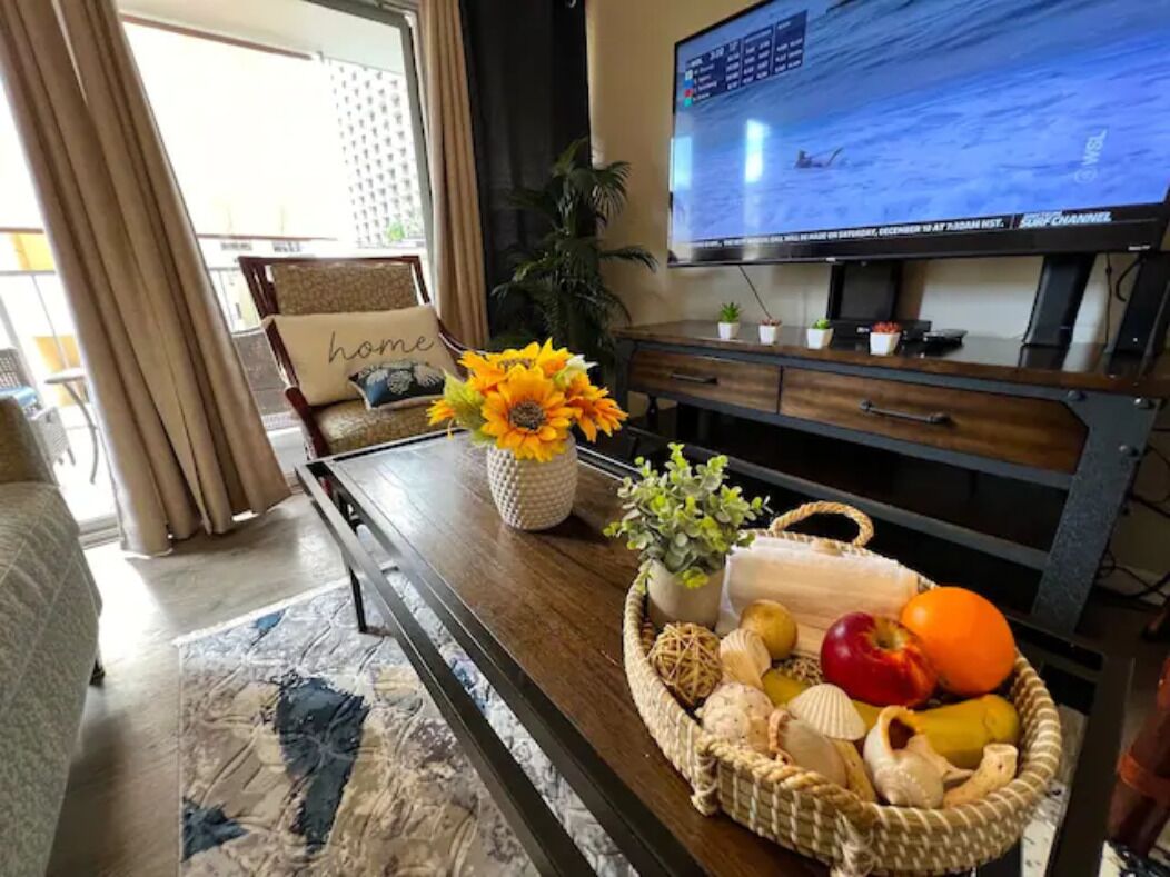 A living room with a television displaying a sports channel, a sofa with a 'home' pillow, a coffee table with a basket of fruits and a vase of sunflowers.