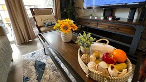 A living room with a television displaying a sports channel, a sofa with a 'home' pillow, a coffee table with a basket of fruits and a vase of sunflowers. - 3 Minute walk to famous Waikiki beach 100% legal with Free Parking  (Honolulu)