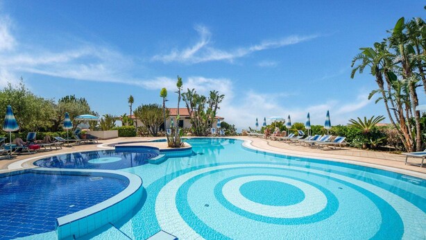 A large, multi-tiered swimming pool with clear blue water, surrounded by lounge chairs and palm trees.