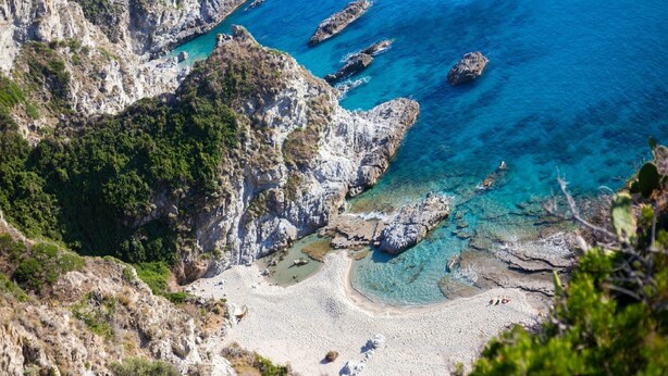 A coastal landscape with rocky cliffs, clear blue waters, and a sandy beach.