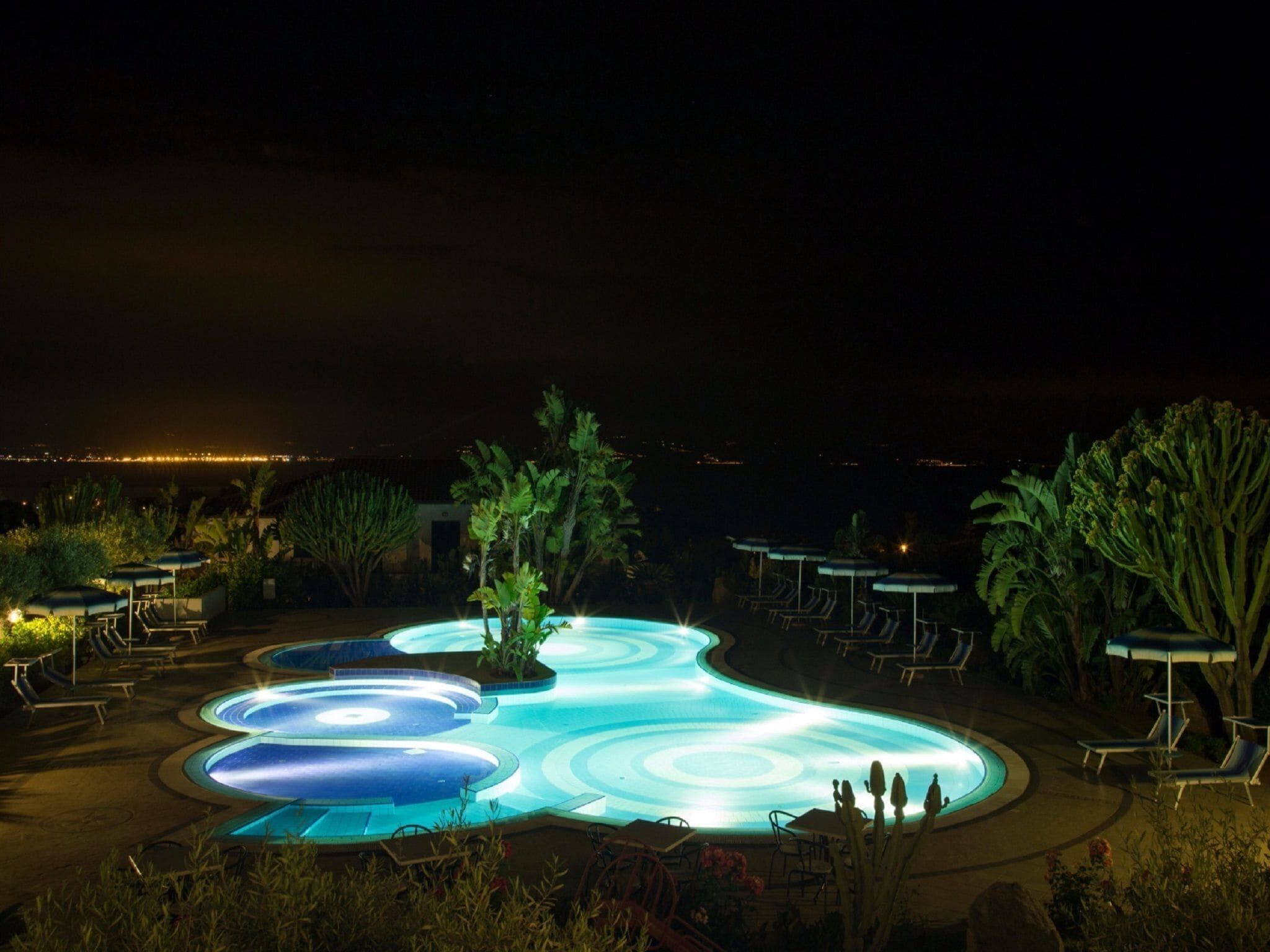 A nighttime swimming pool with a unique design, surrounded by lounge chairs and umbrellas, with a view of the city in the background.