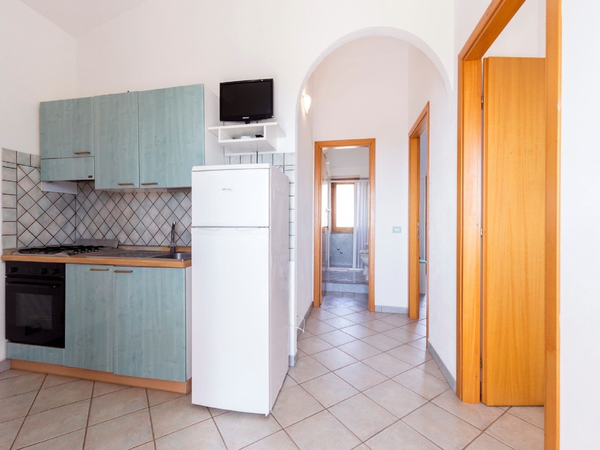Eine kompakte Küche mit türkisfarbenen Schränken, einem weißen Kühlschrank, einem eingebauten Backofen und einem Flachbildfernseher.
