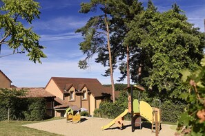 A playground with climbing structures and a slide, surrounded by trees and a building with a tiled roof.