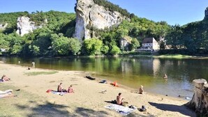 Een zandstrand aan de rivier met mensen die zonnebaden en zwemmen, omringd door groen en een opvallende klif.