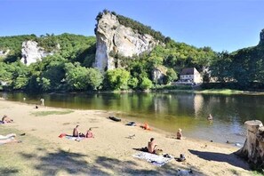 Ein Flussstrand mit Menschen, die sonnenbaden und schwimmen, umgeben von viel Grün und einer markanten Klippe.