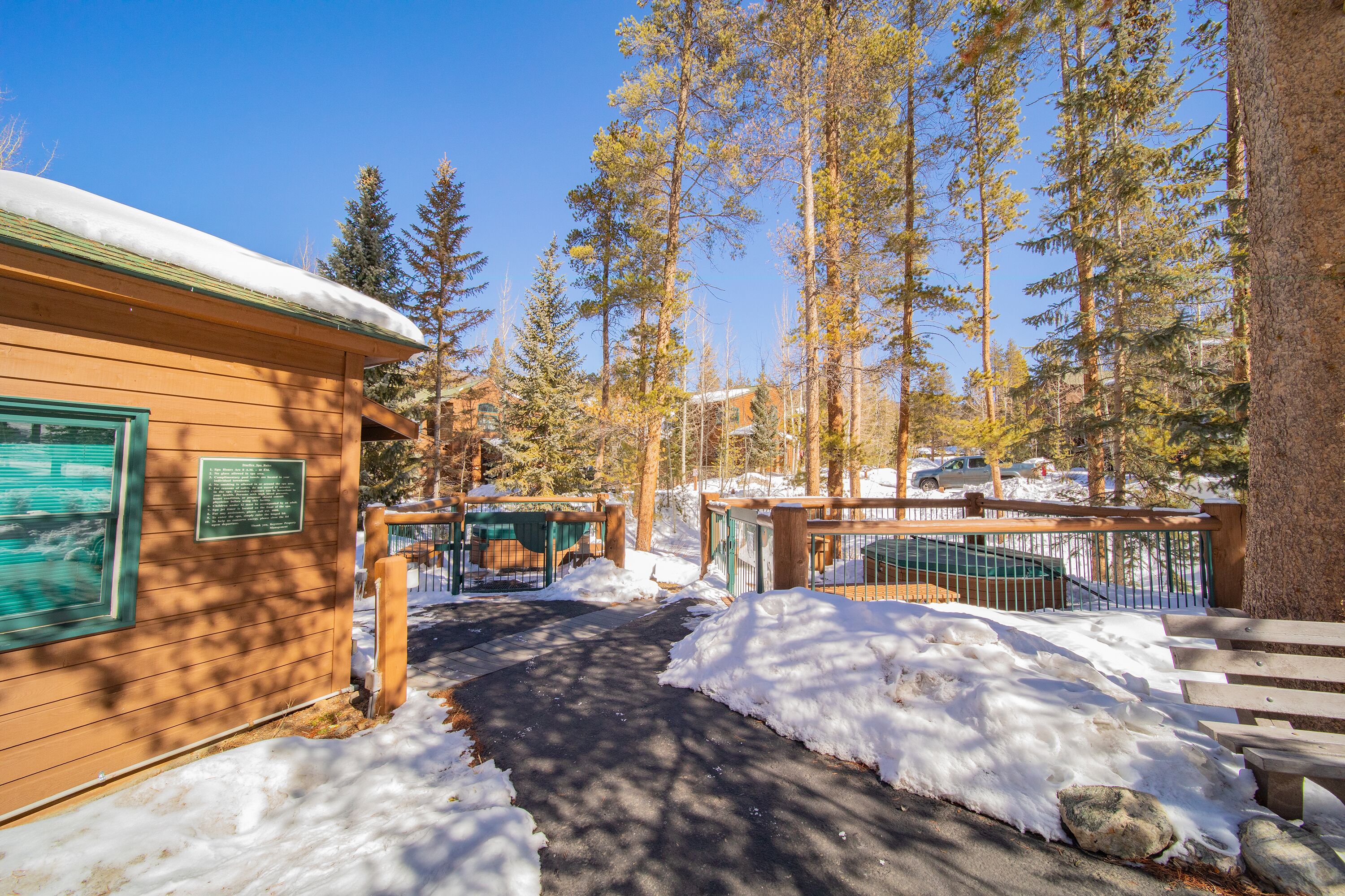 A cabin with a snow-covered roof, surrounded by tall pine trees and a clear blue sky.