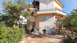 A two-story building with a balcony, a staircase leading to the entrance, and a garden area with potted plants.