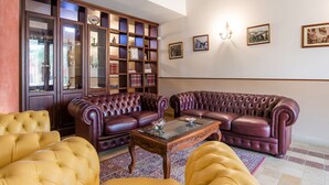 Een woonkamer met een donkerbruine leren bank, een gele fauteuil en een salontafel met een glazen blad. Er staat een houten boekenkast vol boeken en decoratieve objecten.