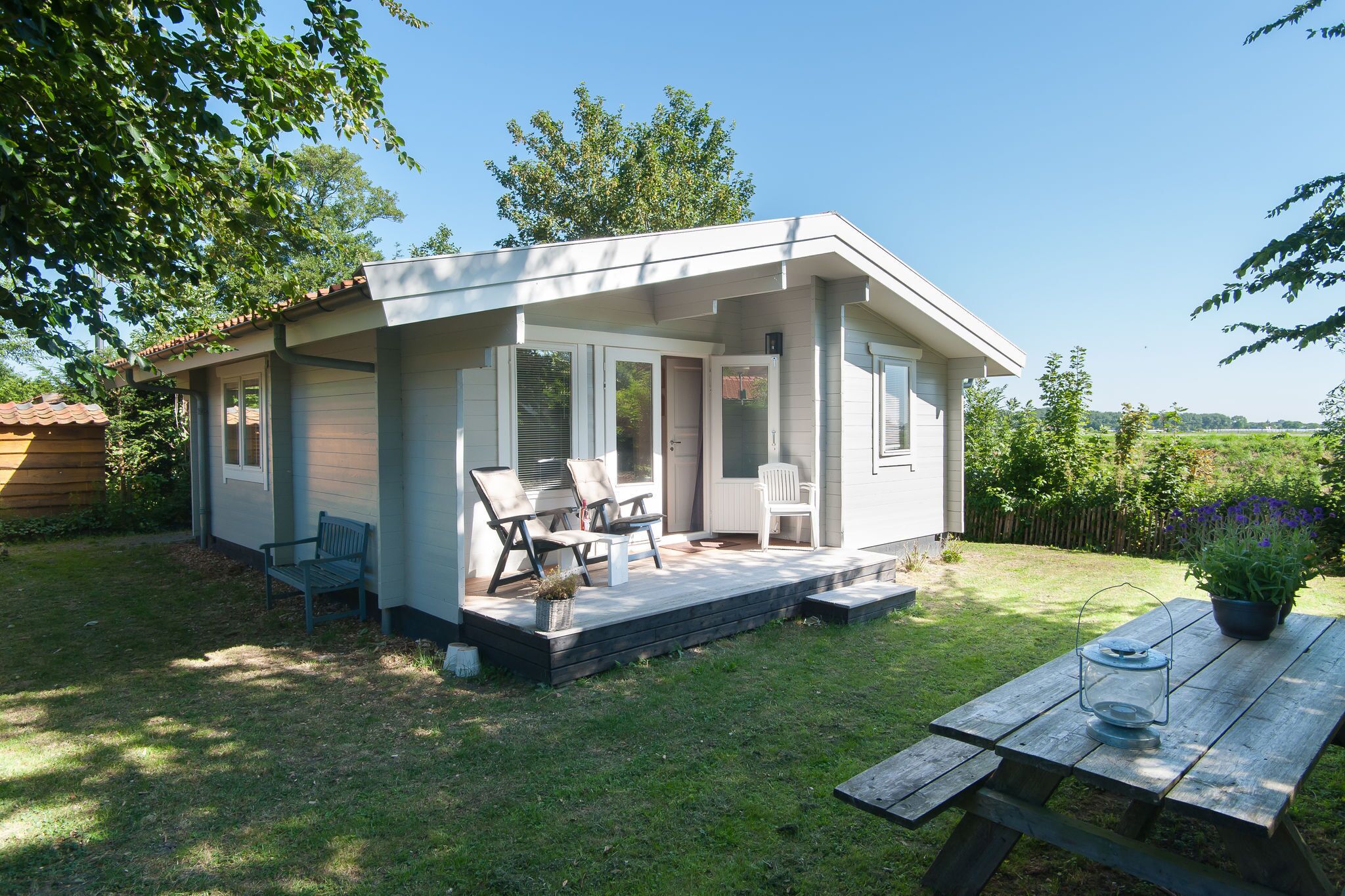 A small, single-story house with a porch, a patio with outdoor furniture, and a wooden table with a lantern.
