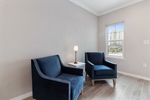 A room with two dark blue upholstered chairs, a small white side table with a lamp, and a large window offering a view of buildings and a beach.