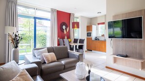A modern living room with a sofa, coffee table, and a TV. There is a kitchen area with a microwave and a TV. The room has large windows and a view of greenery outside.