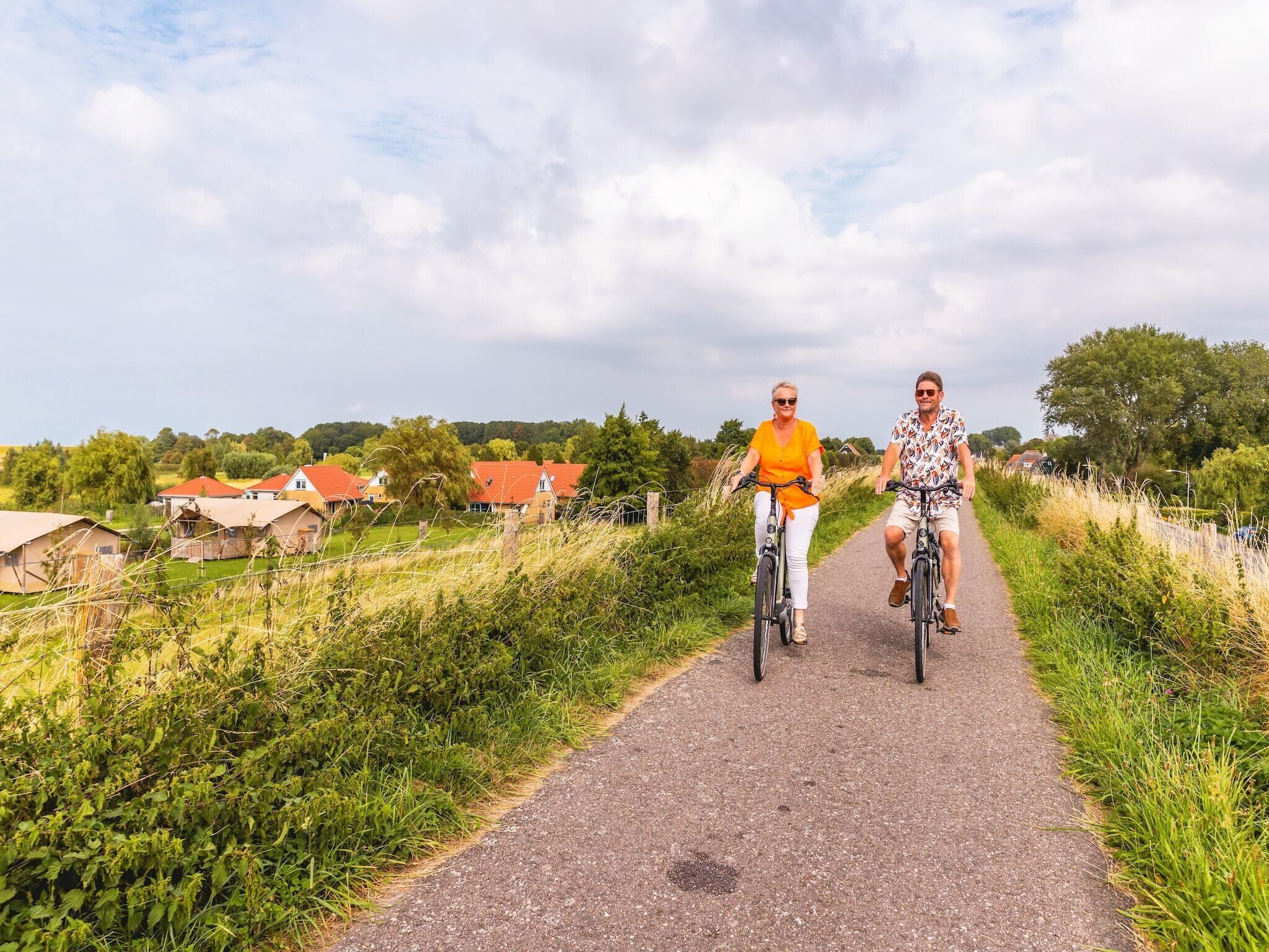 A paved path with two people riding bicycles, surrounded by greenery and houses with red roofs.