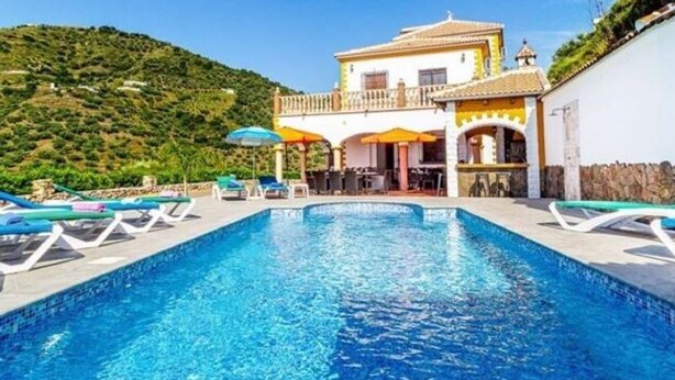 A pool area with lounge chairs and umbrellas, a building with a balcony, and a hillside in the background.