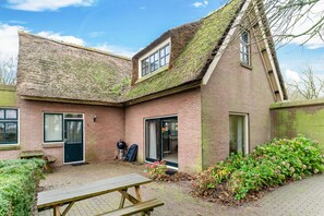 A brick building with a sloped roof covered in moss, a wooden bench, and a small garden area with shrubs and flowers.