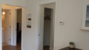 Un couloir aux murs blancs, doté d’un plancher en bois et d’une porte donnant sur un placard équipé d’une tringle à vêtements. Des plantes en pot et un thermostat sont fixés au mur.