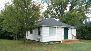 Une petite maison mobile d’un seul étage, avec une porte et des fenêtres vertes, entourée d’une cour verdoyante et d’arbres.