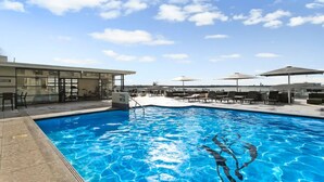 A swimming pool with lounge chairs and umbrellas, a building with glass doors, and a clear sky.