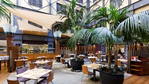 An indoor dining area with tables, chairs, potted plants, and a kitchen in the background.