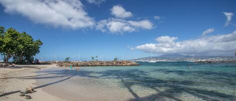 Uma praia com águas claras, rochas e veleiros ao longe.