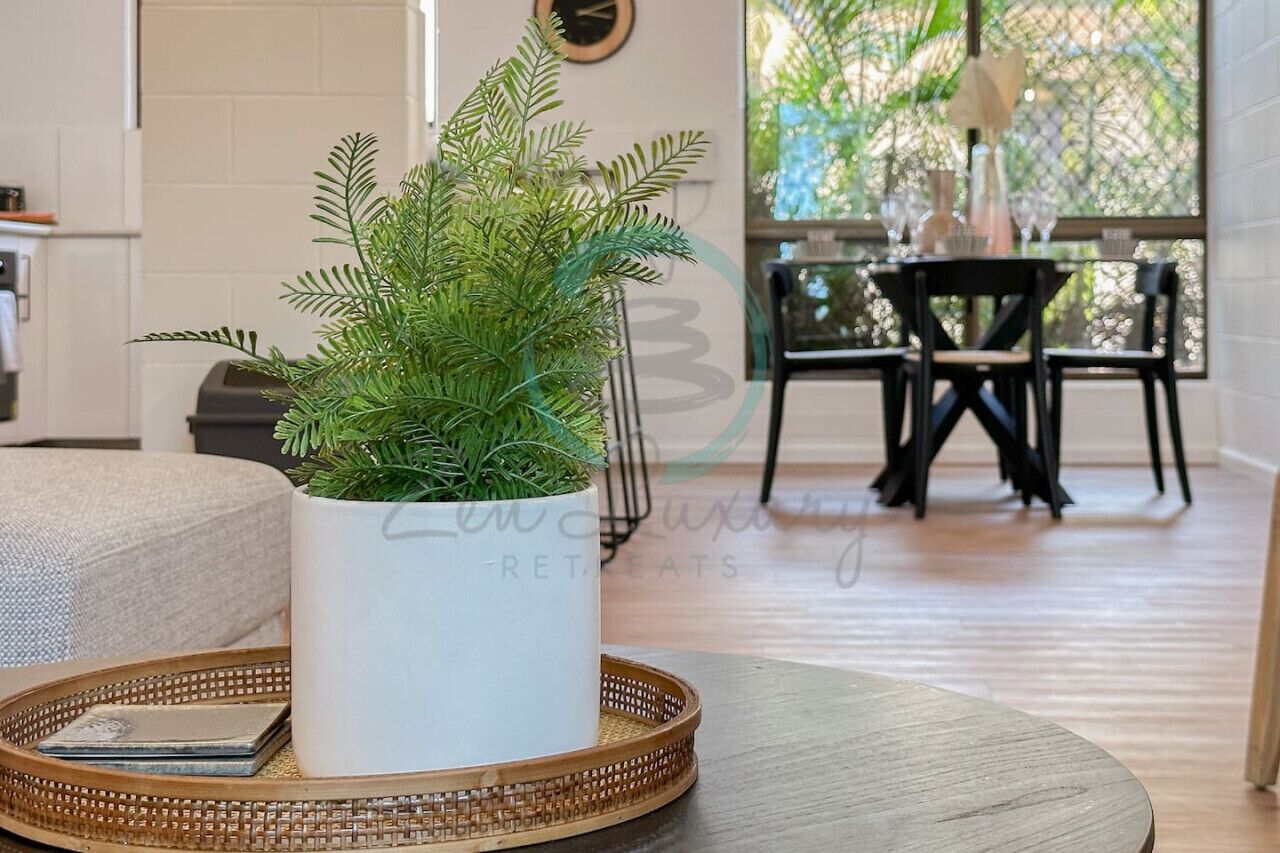 Un salon avec une plante en pot blanche posée sur une table ronde, un plateau en osier, et un coin repas à l’arrière-plan.