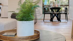 Ein Wohnzimmer mit einer weißen Topfpflanze auf einem runden Tisch, einem Korbtablett und einem Essbereich im Hintergrund.