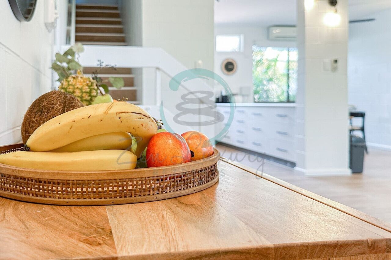 Un panier en osier rempli de fruits est posé sur une table en bois dans une cuisine moderne, avec des meubles blancs et un escalier apparent.