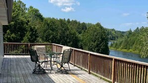 Una terraza de madera con mobiliario exterior, barandilla y vistas a un río y un bosque.