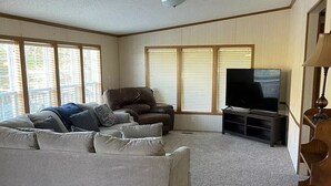 Amplia sala de estar con un sofá gris, un sillón marrón y un televisor sobre un mueble bajo. Ventanas grandes con persianas permiten la entrada de luz natural.