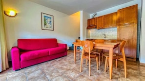 A compact living space with a red sofa, wooden dining table, and matching chairs. A kitchen area with wooden cabinets and a sink is visible.