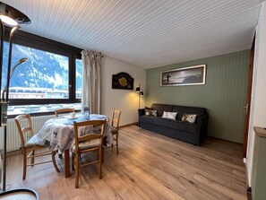 A cozy room with a dining table, chairs, a sofa, a framed picture, and a window with a view of mountains.