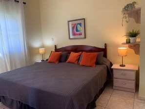 A neatly made bed with a dark bedspread and orange pillows, a bedside table with a lamp, and a wall-mounted shelf with a plant.