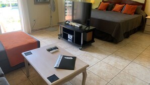 Una habitación de hotel con una cama, un televisor sobre un mueble bajo, una mesa con un libro de bienvenida y una silla.