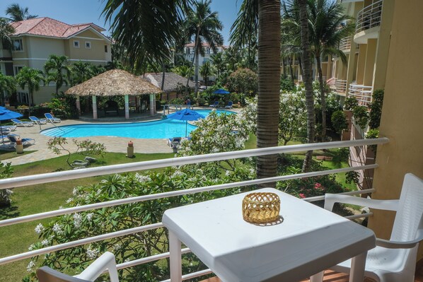 A balcony with a view of a swimming pool, lounge chairs, and a thatched-roof structure.