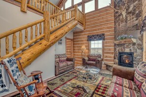 A log cabin interior with a wooden staircase, leather sofas, a glass coffee table, and a stone fireplace. - Rustic Retreat: Unplug and Unwind (Mack)