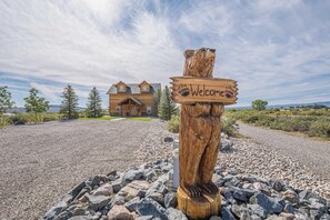 A wooden bear sign with "Welcome" carved on it, standing in front of a house with a gravel driveway and a clear sky. - Rustic Retreat: Unplug and Unwind (Mack)