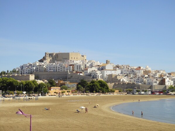 Une ville côtière qui se distingue par une imposante construction en forme de château, située sur une colline, ainsi que par sa plage de sable et son ciel d’un bleu éclatant.