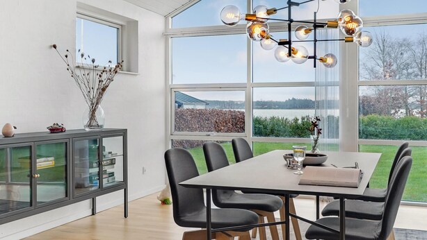 A modern dining area with a large table, black chairs, a glass cabinet, and a view of a lake through large windows. - Gorgeous home in Sunds with kitchen (Sunds)