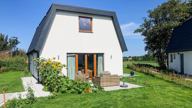 Een modern huis met een schuin dak, een grote glazen deur naar een terras, en een verzorgde tuin met bloemen en gazon.