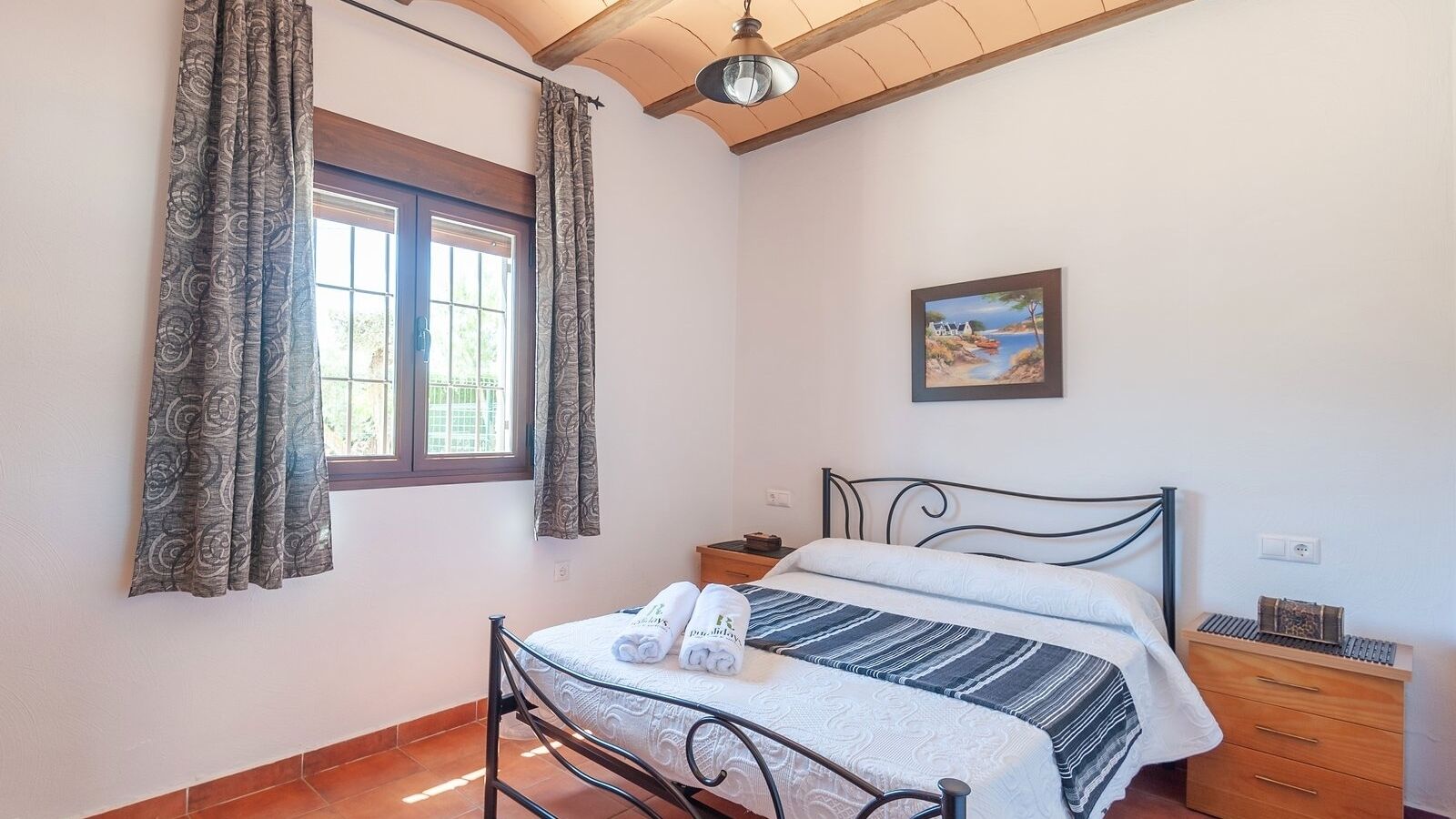 A bedroom with a bed, a window with curtains, a nightstand, a drawer, and a painting on the wall.