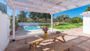 Zona exterior coberta amb una piscina, una taula de fusta i un botic amb flors, i un prat verdós.