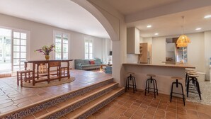 Zona d'estar àmplia amb taula de dinar de fusta, sofà i cuina amb tamboret.