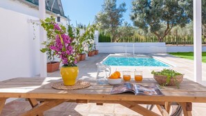 Taula de fusta amb un vas groc de flors, un bol d'albercocs, dues copes de suc d'aranja i una revista. Al fons, hi ha una piscina i plantes en testos.