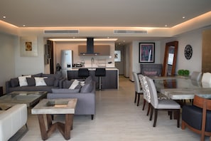 A modern living room with a grey sofa, a glass coffee table, and a dining area with a glass table and chairs. - Beautiful Beach front Condo with pool and Espectacular ocean views (Cruz de Huanacaxtle)