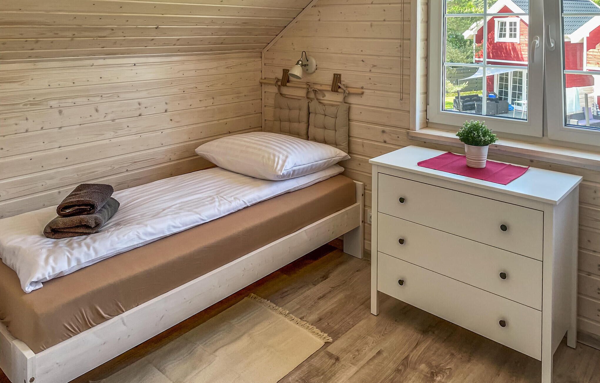 A small, single-bed room with a wooden sloped ceiling, a bedside table with drawers, a window with a view of red buildings, and a small potted plant on the table.