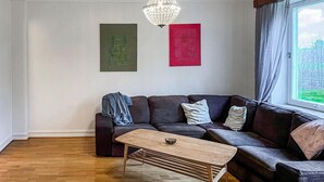 A living room with a dark brown sectional sofa, a wooden coffee table, and a chandelier. There are two framed artworks on the wall.