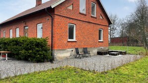 A red brick house with a sloped roof, a gravel driveway, and a small patio with two chairs and a table.
