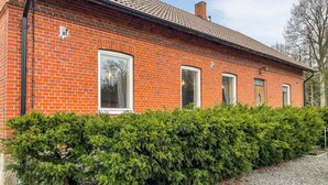 A red brick building with a tiled roof, a large window, and a hedge in front.