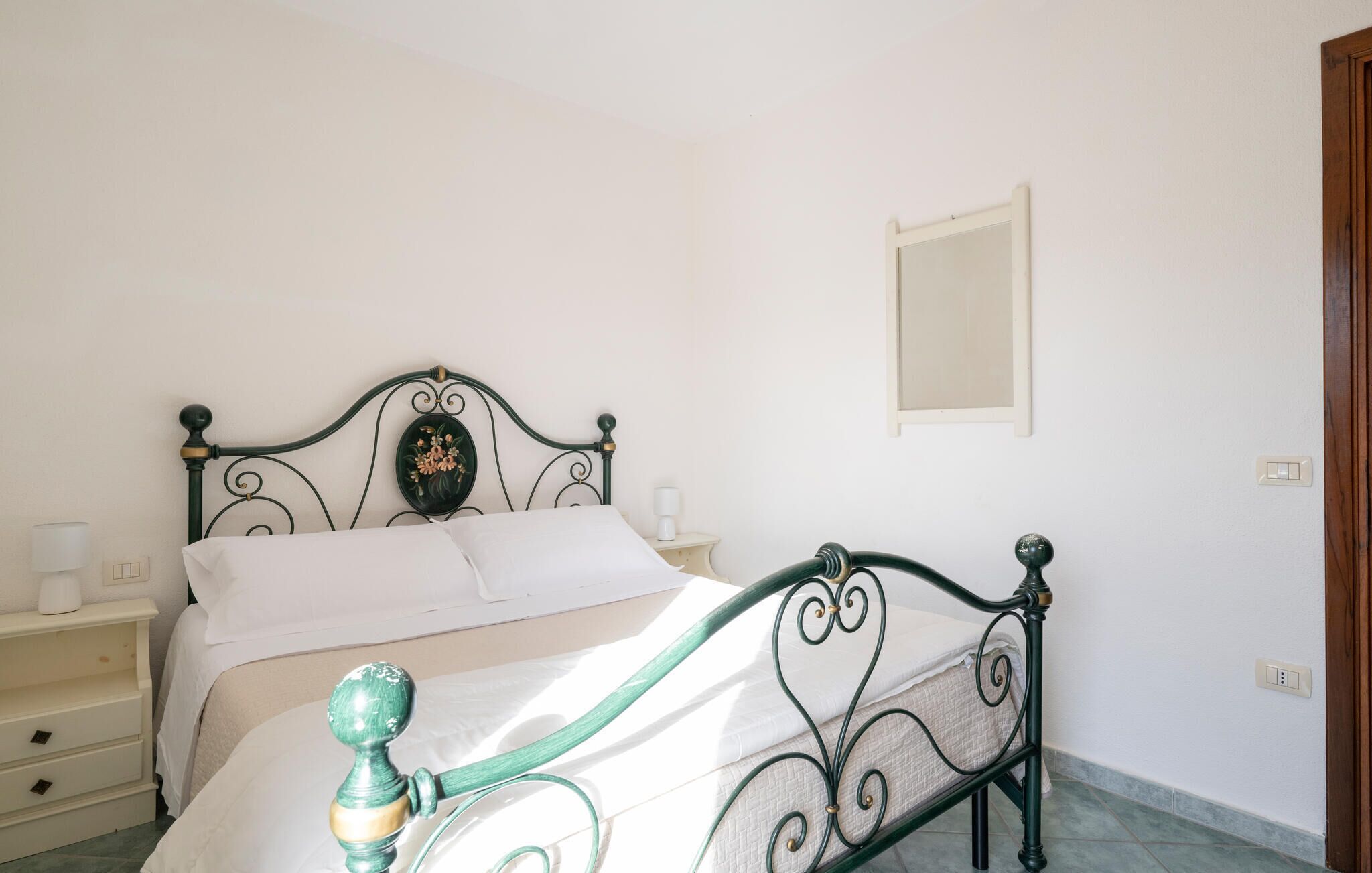 Un letto con struttura in metallo, biancheria bianca e una testiera decorativa. È presente un comodino con una lampada e un piccolo oggetto bianco sopra.